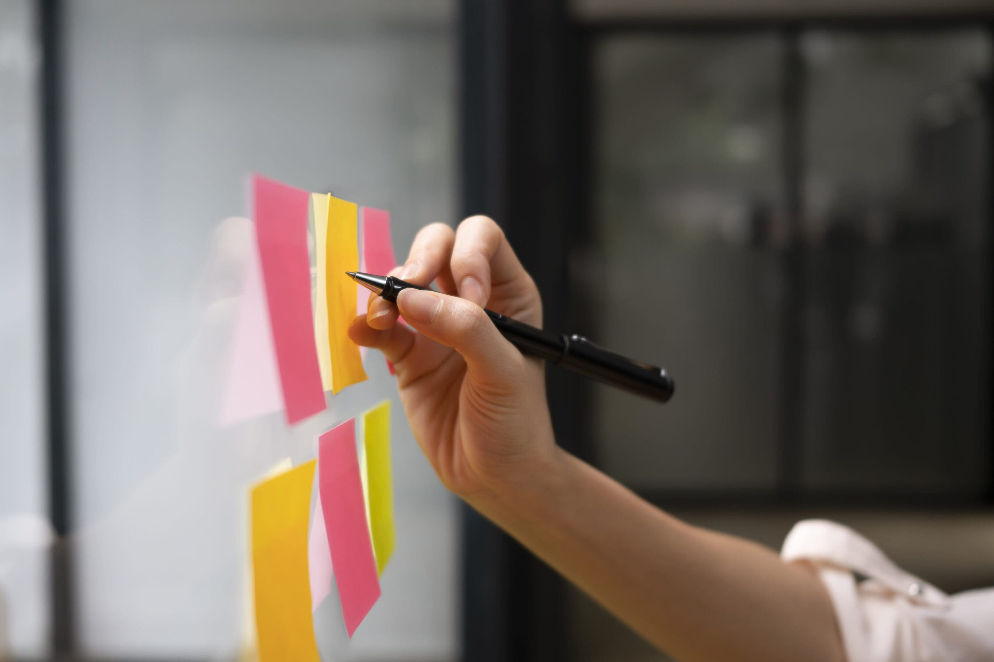 Person writing on colorful sticky notes on a glass wall.