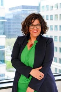 Confident woman in green blouse and navy blazer smiling indoors.